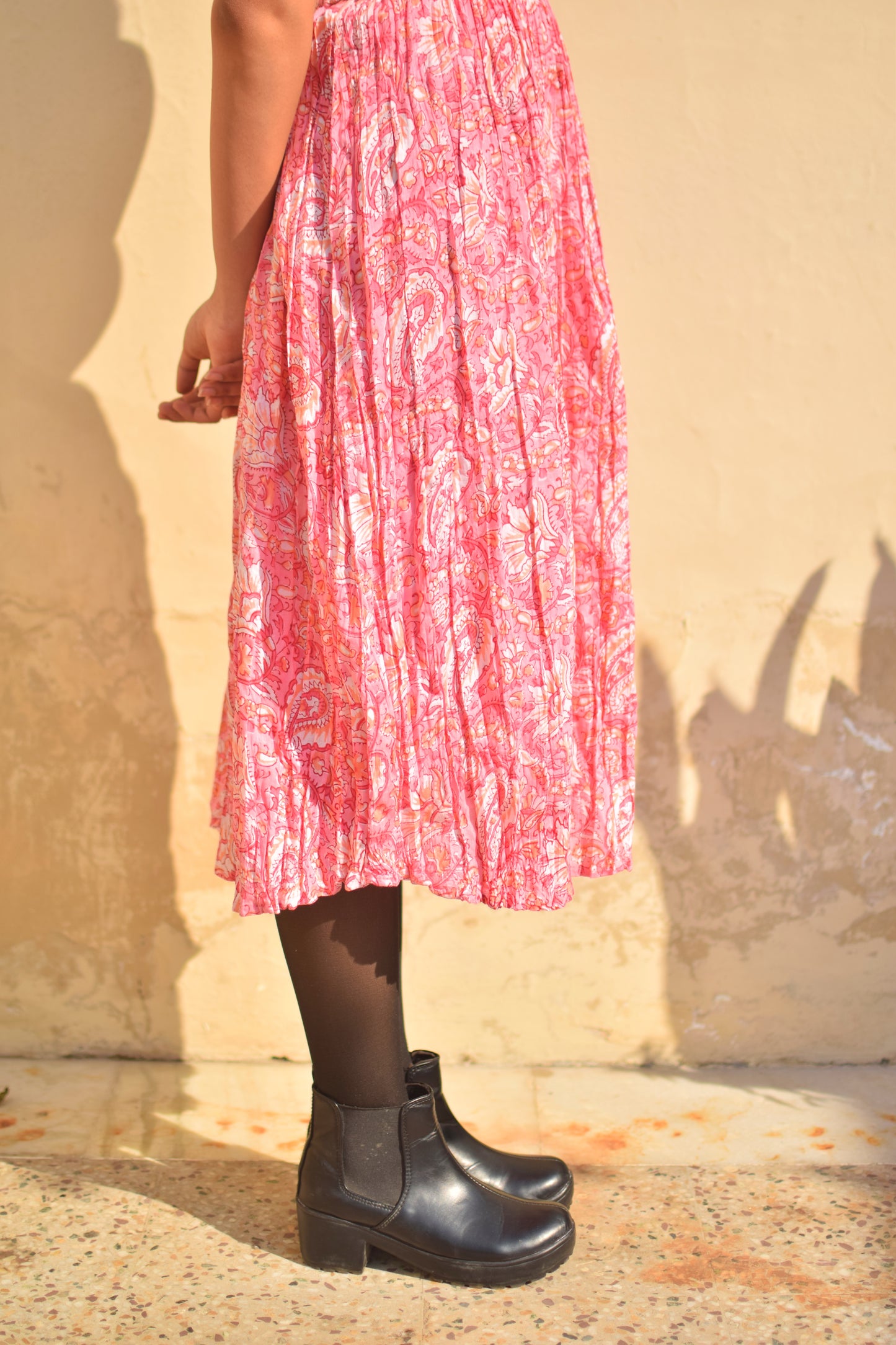 Person wearing a pink patterned dress and black boots against a beige wall.