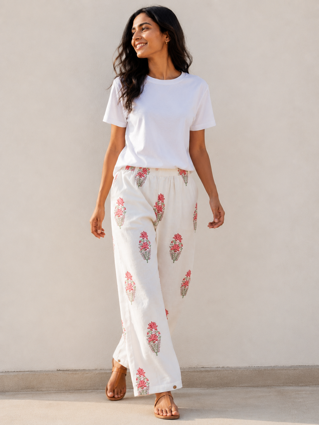 A woman wearing a white t-shirt and white relaxed-leg pants with pink and green floral hand blocked prints, standing against a plain beige wall.