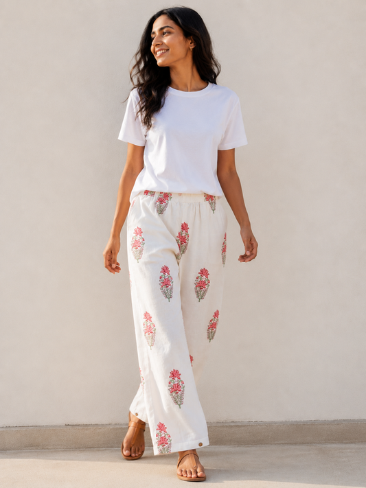 A woman wearing a white t-shirt and white relaxed-leg pants with pink and green floral hand blocked prints, standing against a plain beige wall.