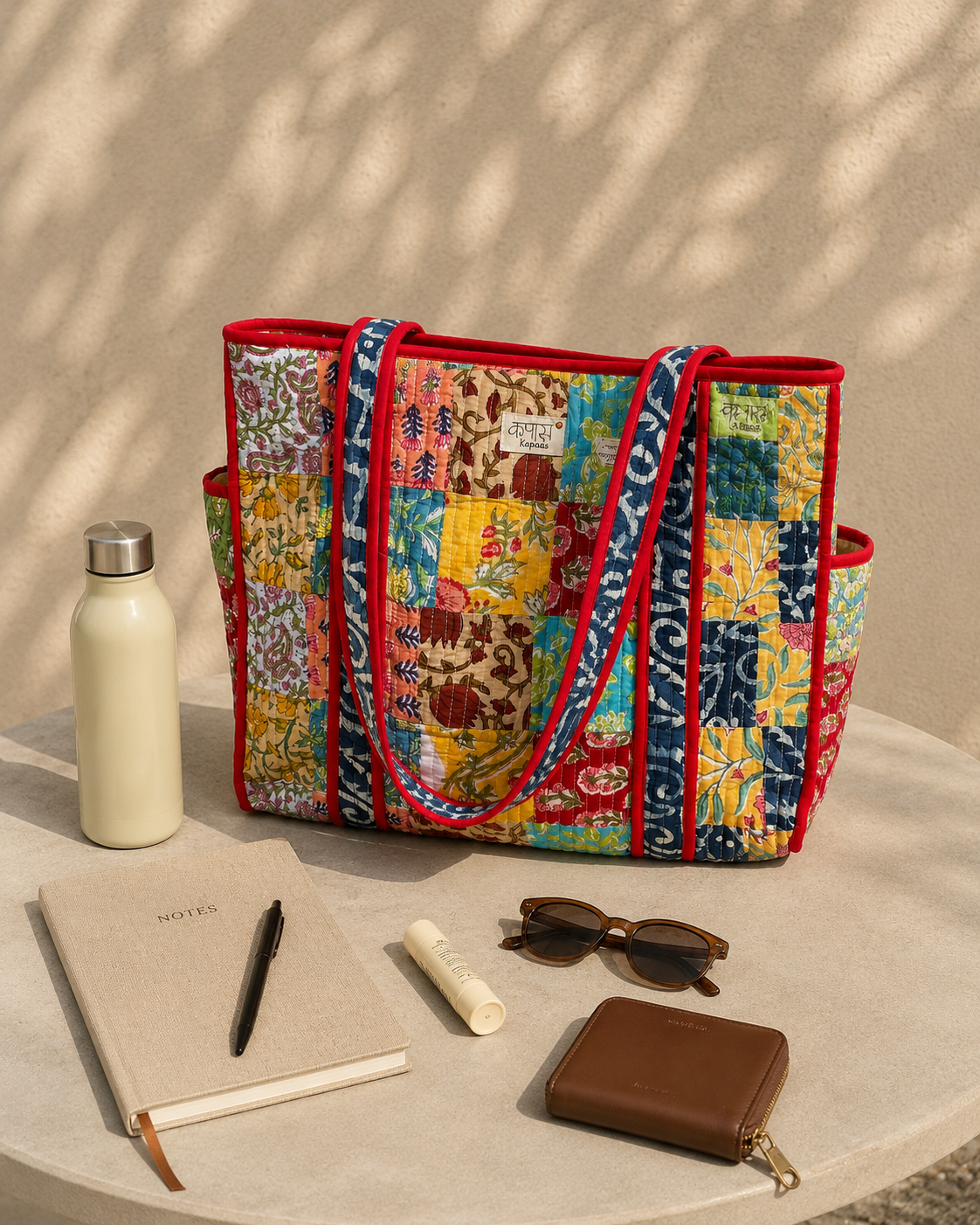 Colorful patchwork tote bag on a table with a notebook, pen, wallet, and sunglasses.