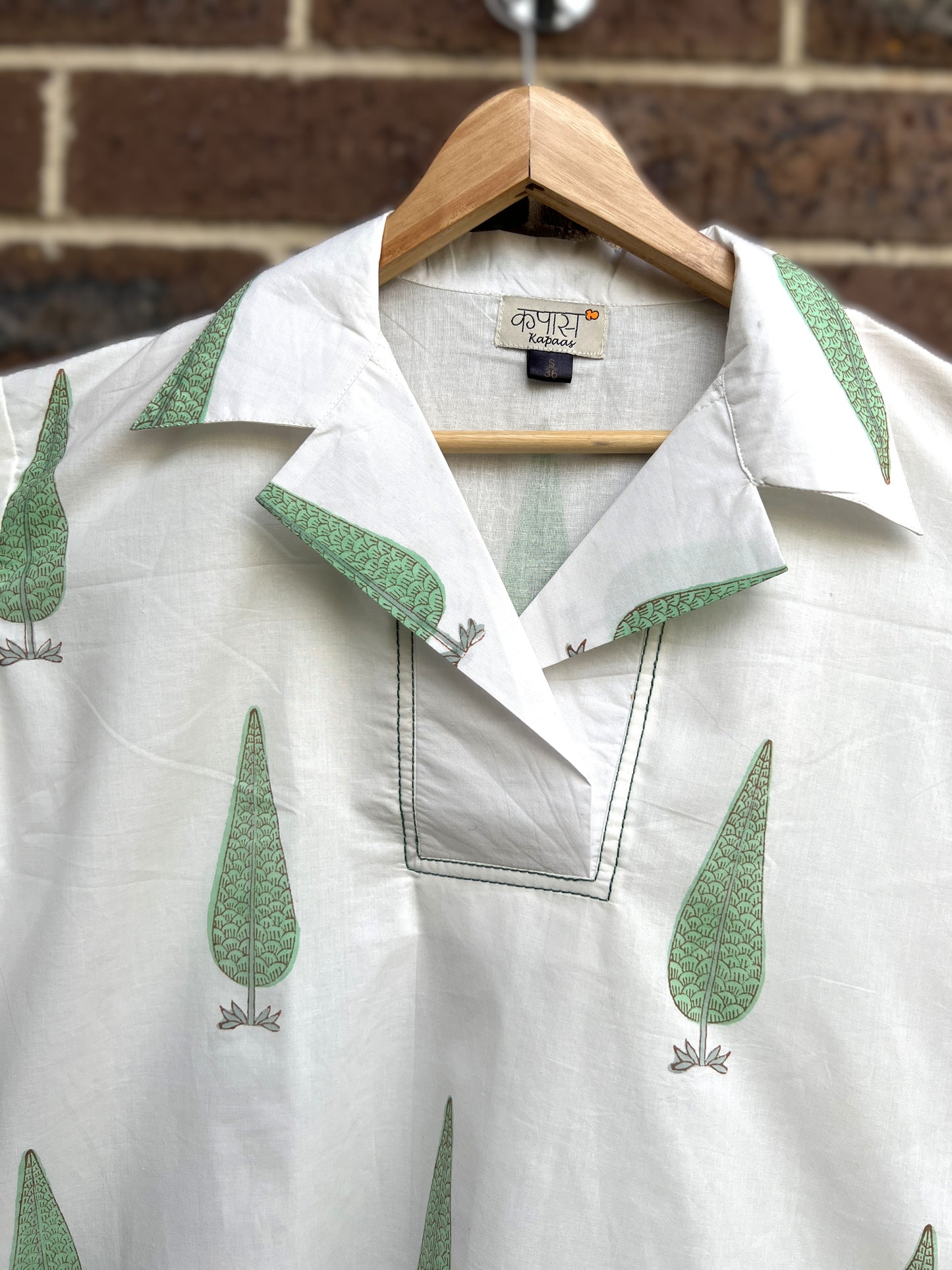 White shirt close up with green leaf patterns on a wooden hanger against a brick wall.