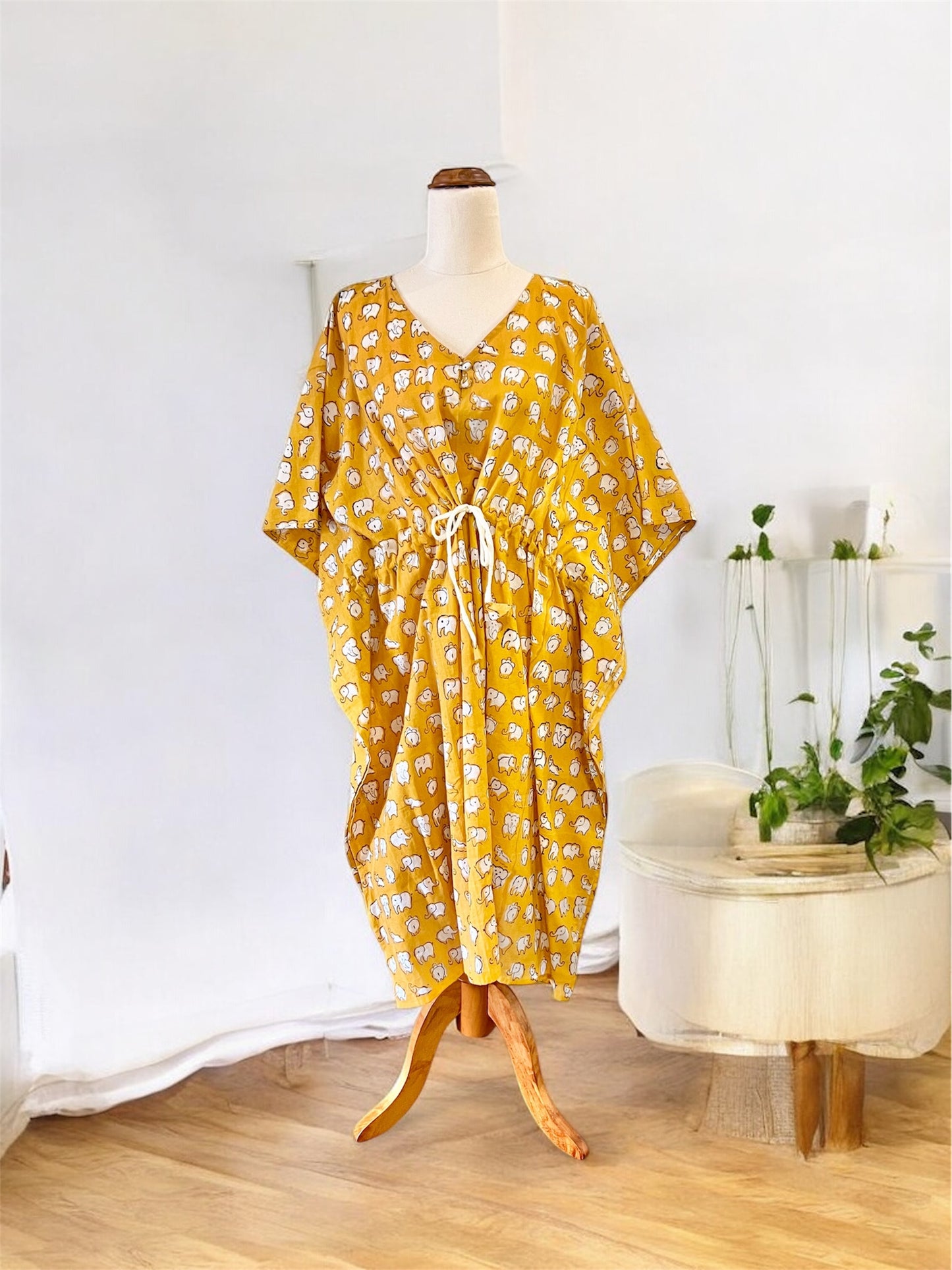Yellow elephant hand block printed kaftan on a mannequin against a white wall with a small table and plants in the background.