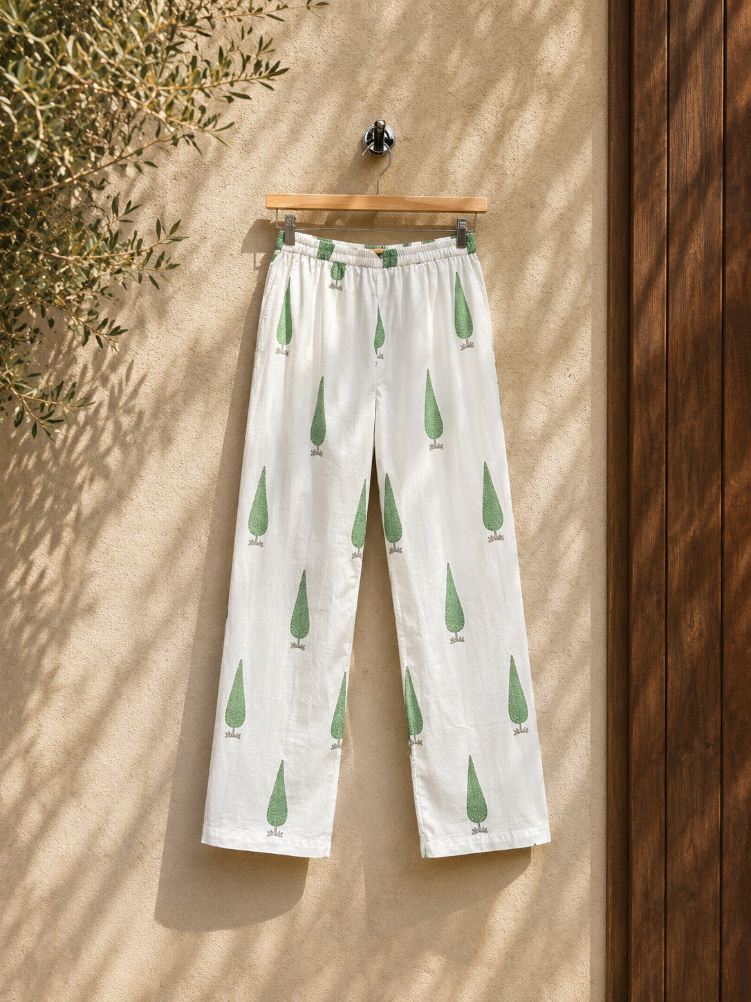 White pants with sage cypress print  hanging on a wooden hanger against a beige wall outdoor.
