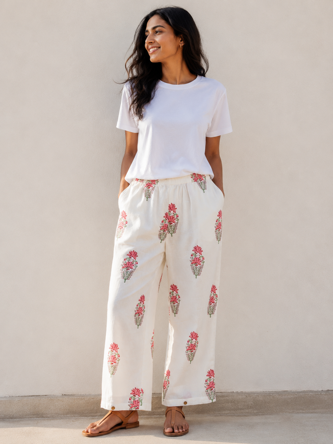 A woman wearing a white t-shirt and white relaxed-leg pants with a pink and green floral hand blocked print, standing against a beige wall.