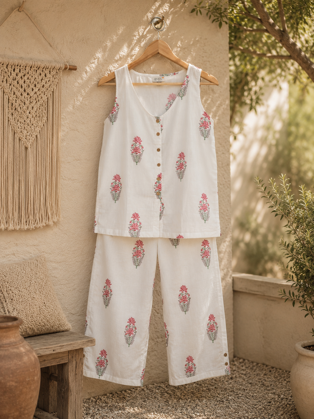 A two-piece sleeveless co-ord set hanging on a wooden hanger against a rustic beige stucco wall. The set features a button-front top and matching relaxed-leg pants, both in an ivory coloured cotton fabric with pink and green floral hand block prints.