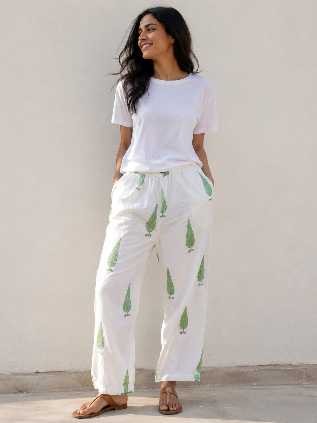Woman wearing a white t-shirt and white sage cypress print pants standing against a plain background outdoor.