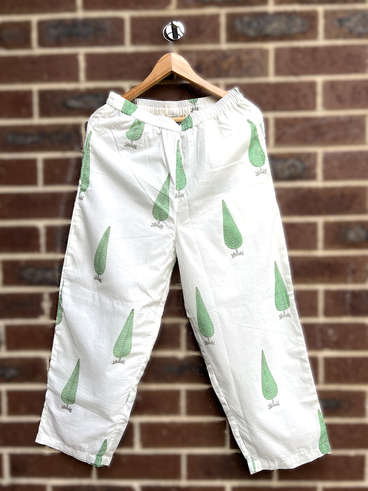 White sage cypress print pants hanging on a wooden hanger against a brick wall.