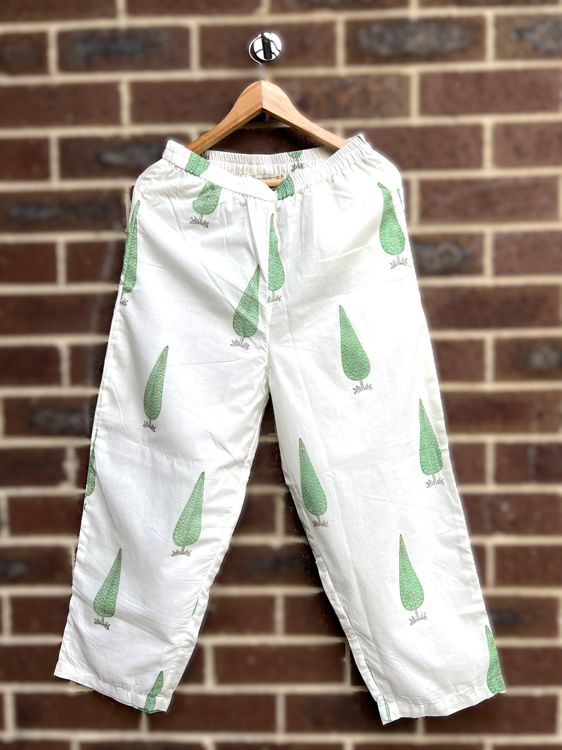 White sage cypress print pants hanging on a wooden hanger against a brick wall.