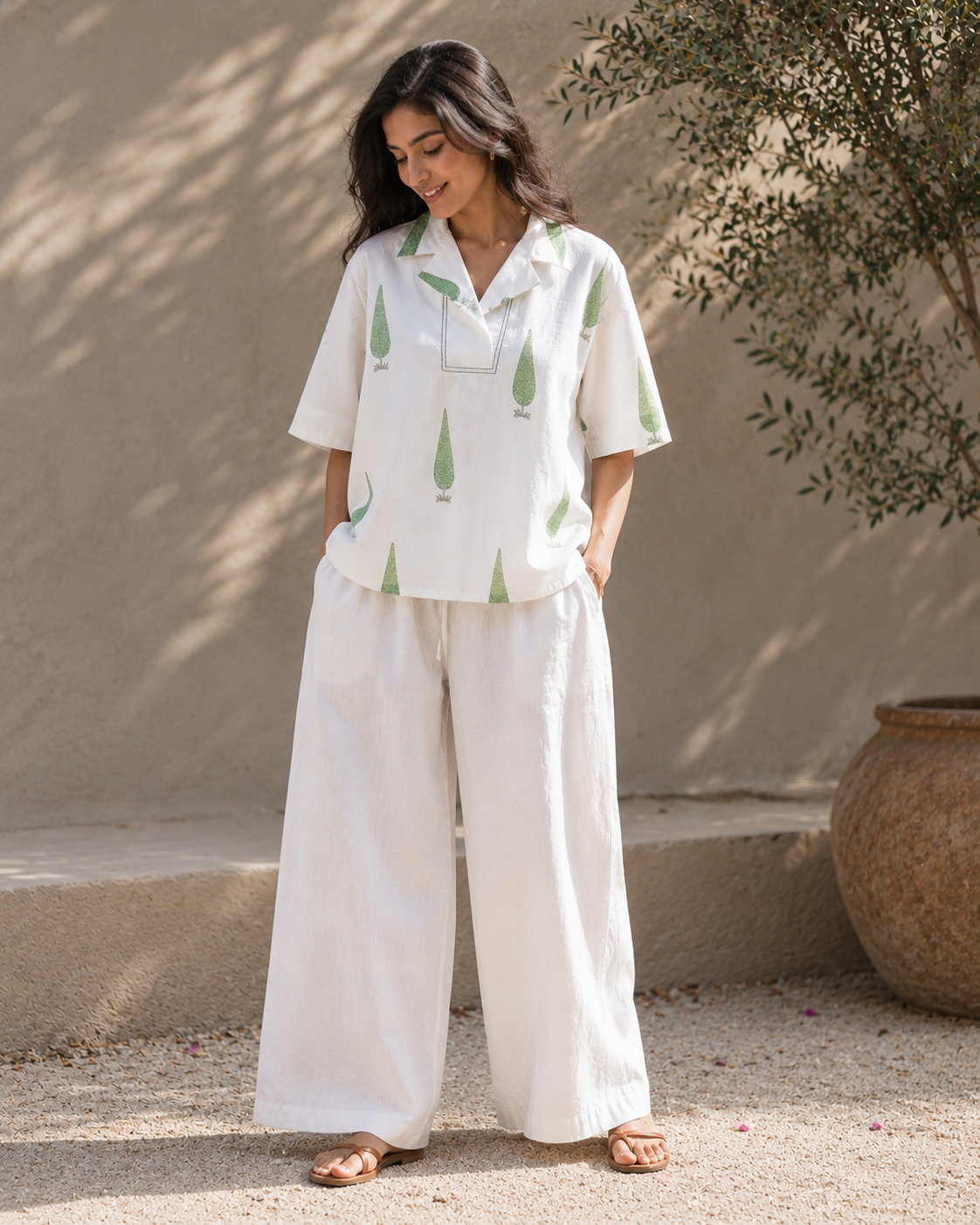 Woman wearing a white sage cypress print lounge shirt and white pants standing against a beige wall.