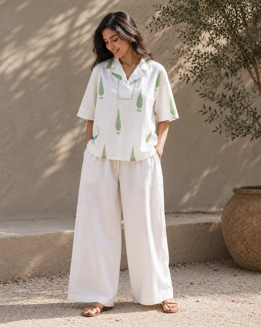 Woman wearing a white sage cypress print lounge shirt and white pants standing against a beige wall.